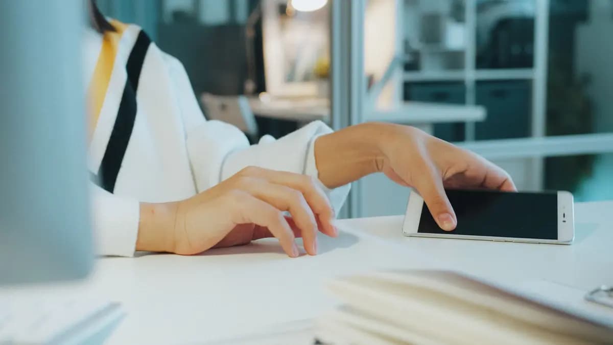 Close-up of a woman's hand using a smartphone at a desk in a modern office.